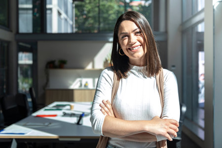 Portrait of smiling young brunette woman looking at camera standing with arms crossed in front of desk in coworking space. Female entrepreneur. Copyspace. Startup concept. Co working space concept.の写真素材