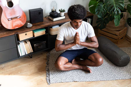 Young african american man meditating with hands in prayer in bright living room. Meditation concept. Spirituality concept.の写真素材