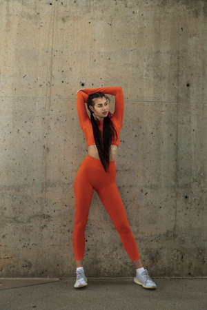 Female athlete wearing orange sportswear resting to stretch triceps. Woman standing workout clothes.