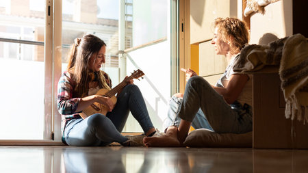 Two women relax after a day of moving houses. Young caucasian woman plays the ukelele. Horizontal...