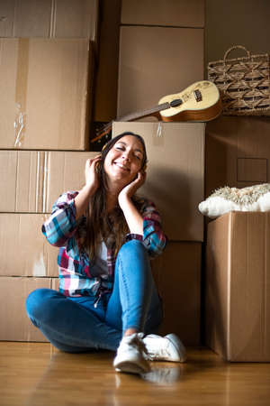 Young caucasian woman listens to music with headphones after a day of moving houses. Pile of cardboard boxes. Vertical.の写真素材