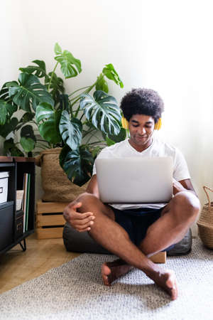 Young african american man using his laptop and yellow headphones in his bright apartment. Vertical image. Copy space.の写真素材