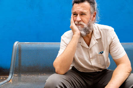 Pensive mature man with hand on face sitting on a bench. Sad and worried expression. Copy space.の写真素材
