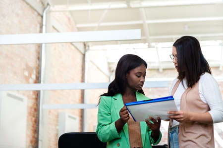 Two multiracial female coworkers stand in the office look over some documents. Copy space.の写真素材
