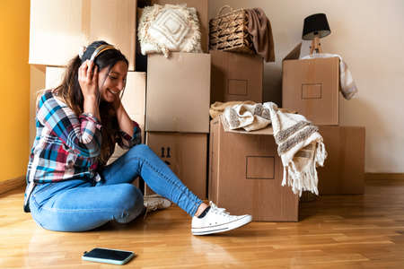 Young woman listens to music with wireless headphones in front of pile of cardboard boxes and decorative house objects.の写真素材