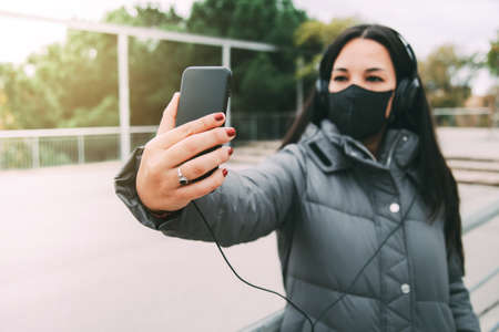 Young asian woman with headphones and face mask taking selfie with mobile phone outdoors.の写真素材