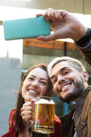 Smiling young couple enjoying a beer together and taking a selfie with mobile phone. Vertical image.