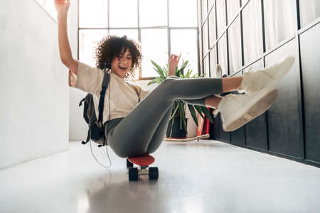 Playful young multiracial woman sliding down a corridor sitting on skateboard having fun.