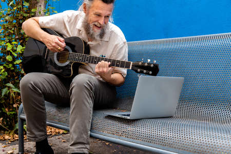 Mature caucasian man sitting on a bench learning to play the guitar with online classes using laptop.の写真素材