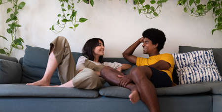 Smiling young interracial couple sitting on the couch enjoy some time together at home talking...