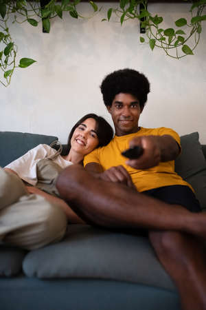 Young interracial couple watch TV at home. African american man changes channel with remote control....