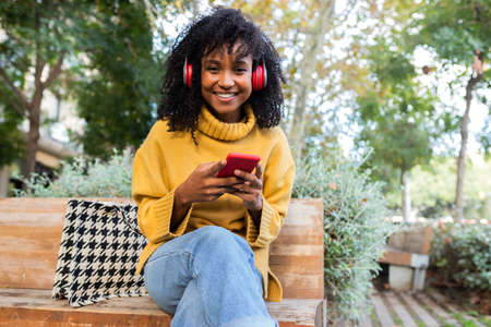 Young African American woman wearing headphones using cellphone sits on park bench looking at...