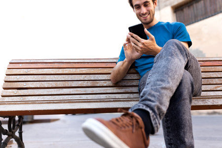 Young happy caucasian man relaxing sitting on a bench using cellphone. Selective focus on hand. Copy space.の写真素材