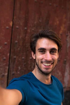 Smiling young caucasian man looking at camera takes selfie with wood background. Vertical.の写真素材
