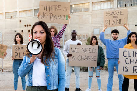 Young woman holds megaphone looking at camera leads demonstration against global warming. Protesters in background.の写真素材