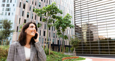 Panoramic image of smiling young business woman talking on cellphone next to office buildings. Copy...