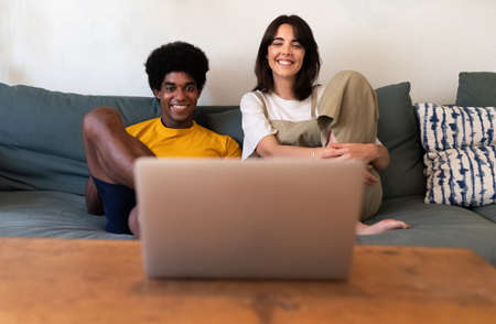 Smiling young multiracial couple on the sofa watching tv show on laptop together.