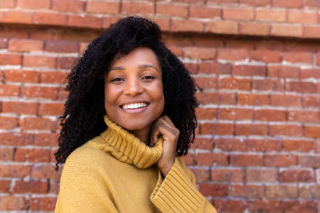 Smiling African american woman looking at camera wearing yellow sweater. Orange brick background. Copy space.の写真素材