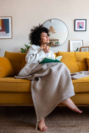 Pensive happy African american woman reading a book at home sitting on sofa relaxing drinking coffee. Vertical image.の写真素材