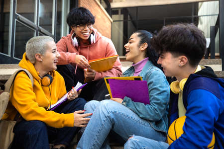 Multiracial group of gen z college students talk and laugh sitting on campus stairs outside. Friends having funの写真素材