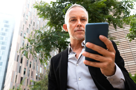 Middle age business man using cell phone to read e-mails outside office buildings. Copy space.