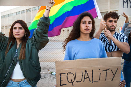 Multiracial people march together protesting for LGBT rights holding cardboard banners, waving...
