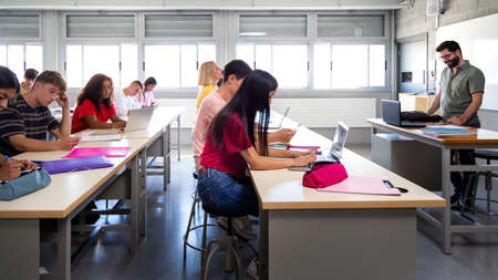 Side view of group of multiracial students studying and using laptops in class with male teacher.の写真素材