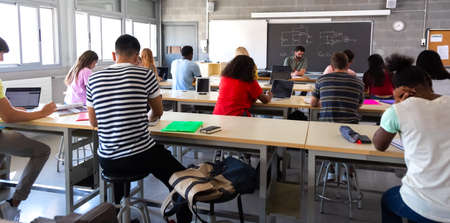 Rear view of group of multiracial high school students in class using laptops while teacher marks exams.の写真素材