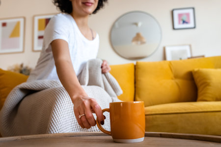 Unrecognizable woman hand grabs cup of coffee. Woman having tea sitting on the couch at home. Copy space.の写真素材