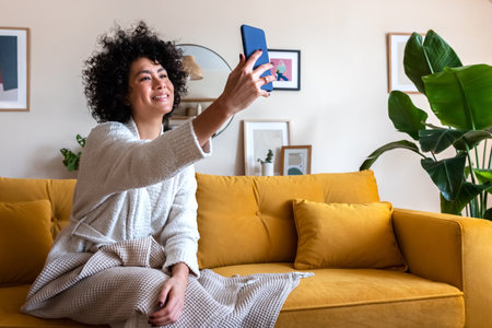 Smiling African american woman sitting on couch taking selfie photo at home using mobile phone.の写真素材