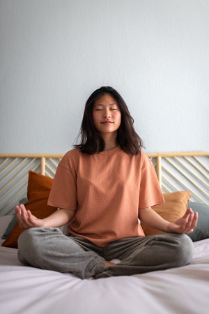 Front view of college student woman meditating and doing breathing exercises on bed in the morning. Verticalの写真素材