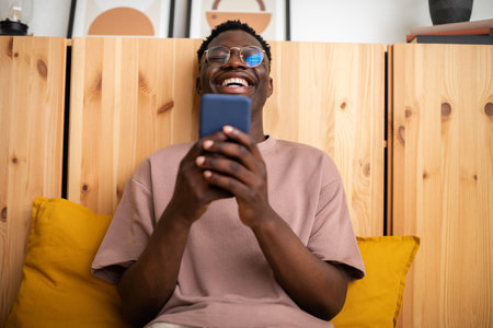 Young black man laughing at home relaxing using mobile phone to watch funny videos. Reading text message.の写真素材