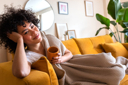 Happy African American woman relaxing at home lying on couch holding cup of warm coffee looking at camera. Copy space.の写真素材