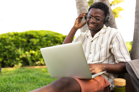 Black man listens to music with headphones and using laptop outdoors. Student doing homework on campus. Copy space.の写真素材