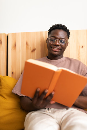 Vertical portrait of young African American man with glasses relaxing at home, reading a book.