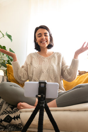 Young caucasian woman influencer vlogger recording herself with smartphone at home. Live streaming...