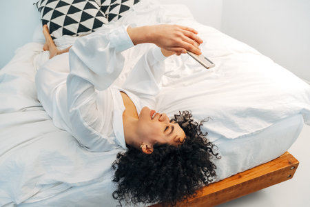 Young mixed race woman looking at cellphone browsing through social media lying upside down on the bed. Copy space.の写真素材