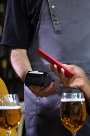 Vertical close up of unrecognisable person paying bill in bar with mobile phone. Waiter holding dataphone.の写真素材