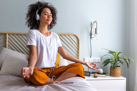 Young woman meditating at home sitting on bed following online meditation, listening with headphones, using phone app.の写真素材