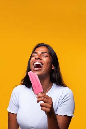 Vertical portrait of Indian woman laughing eating pink ice pop on yellow background. Asian woman...