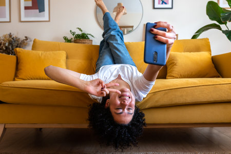 Joyful African American woman lying upside down on the sofa takes selfie with smart phone. Social media.の写真素材