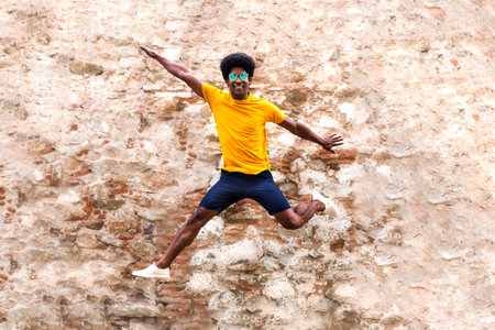 Happy young African American man jumping in the air. Rustic brick and stone wall background.の写真素材