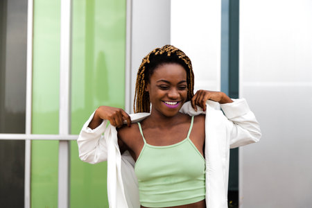 Happy young Black woman with blonde braids putting on jacket outdoors.