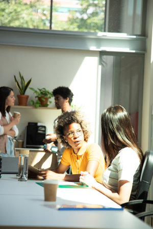 Two female coworkers share ideas while two colleagues have coffee break in the background. Vertical...