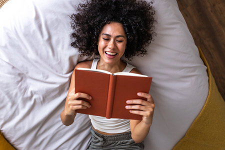 Top view of young happy multiracial woman lying on the bed reading a book laughing.の写真素材
