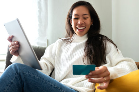 Happy young Asian woman using digital tablet and credit card to shop online sitting on the sofa at...