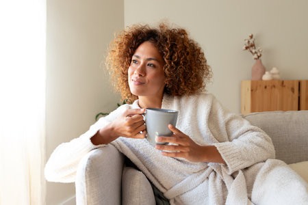 Pensive multiracial woman relaxing at home, sitting on the sofa drinking tea.の写真素材