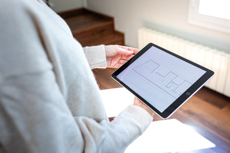 POV of woman holding digital tablet looking at floor plan standing indoors.