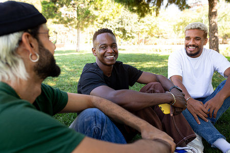Young African American man and his male friends laughing and having fun together sitting on the grass in a park.の写真素材