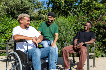 Young disabled African American man in a wheelchair and his male friends talking sitting on a bench in city park.の写真素材